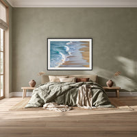 Tura Beach Waves framed print in black with a white mat, above a khaki linen bed against a green-grey wall in a bedroom