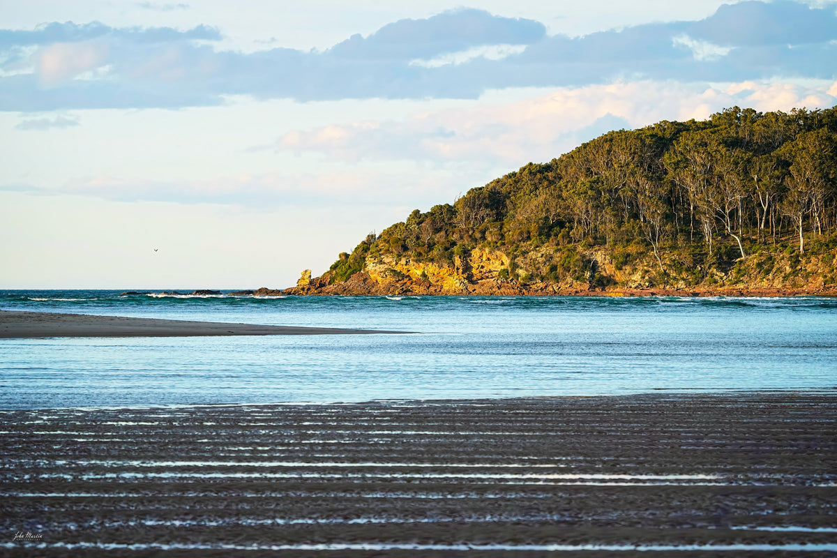 Ribbed wet sand and shallow water at Pambula River mouth below a sunlit forested headland — fine art print