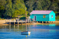 Close view of Mitchies Jetty boatshed, turquoise with a pink roof and yellow window frames, a small sailing boat moored in the foreground — fine art print