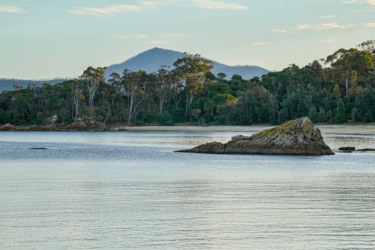 A rocky outcrop in the calm water of Twofold Bay, Eden, the forested shore and a mountain peak beyond — fine art print