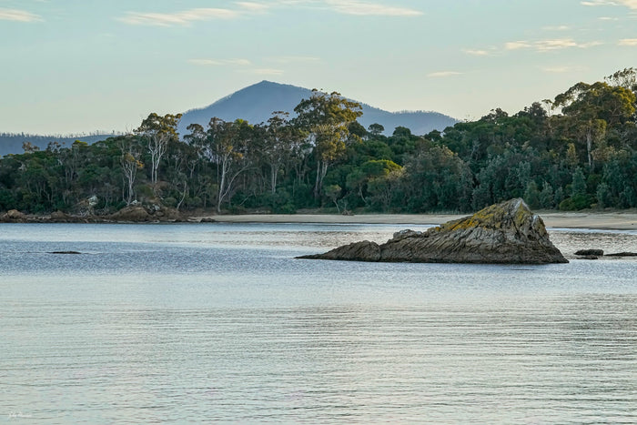 A rocky outcrop in the calm water of Twofold Bay, Eden, the forested shore and a mountain peak beyond — fine art print