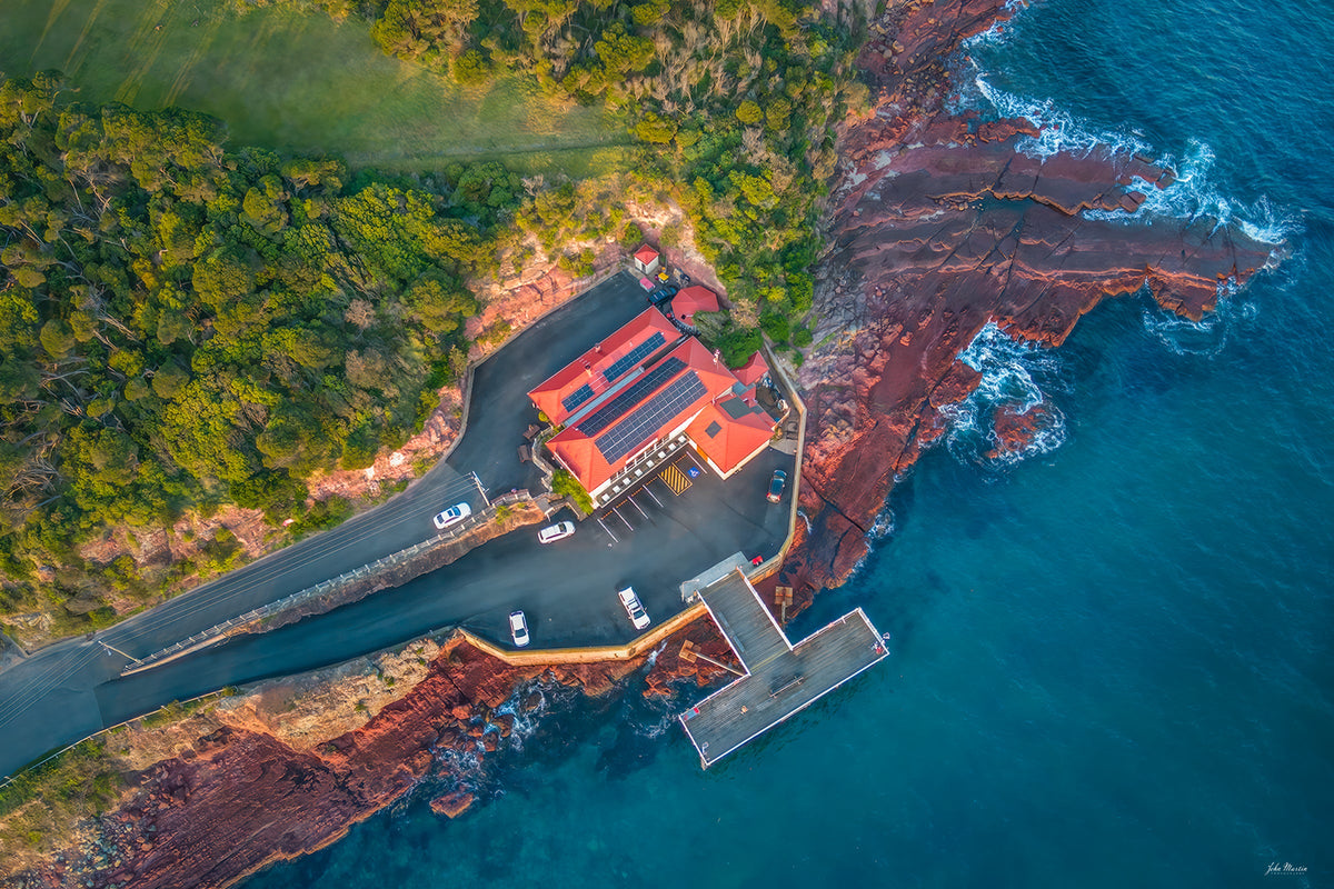 Merimbula Wharf and its red-roofed restaurant seen from directly above, the timber wharf reaching into turquoise water over red rock platforms and breaking surf — fine art print