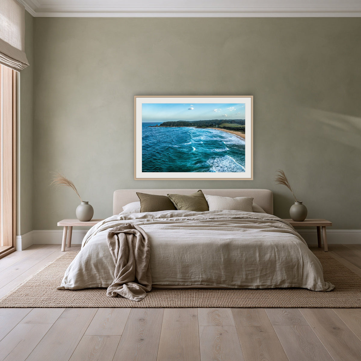 Cuttagee Beach framed print in oak with a white mat, above a linen bed against a sage wall in a bedroom