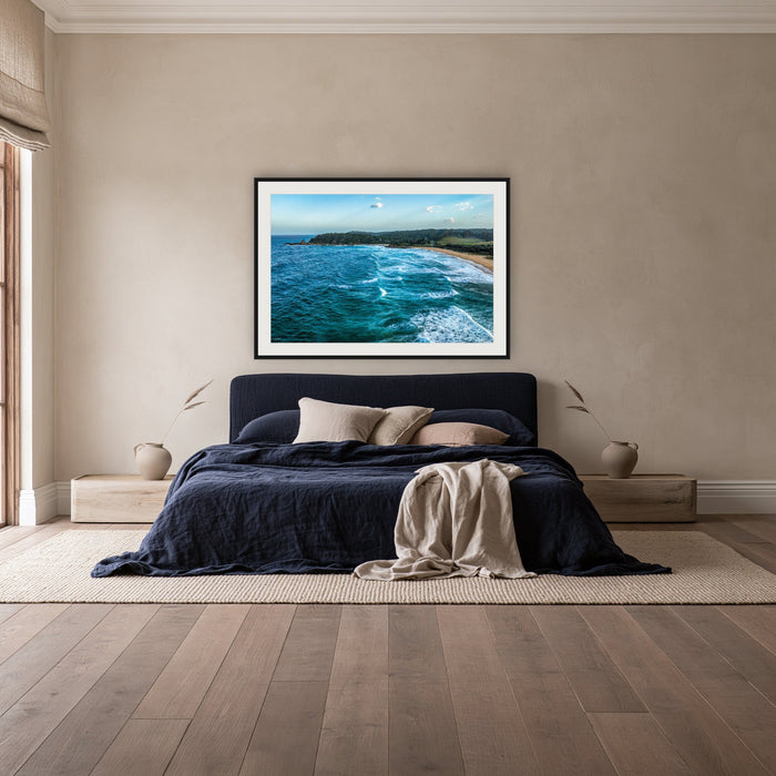 Cuttagee Beach framed print in black with a white mat, above a navy linen bed in a bedroom