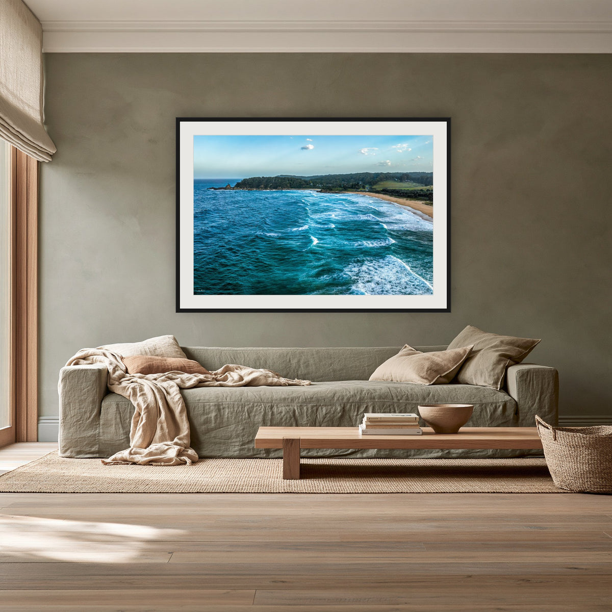 Cuttagee Beach framed print in black with a white mat, above a sage linen sofa in a lounge room