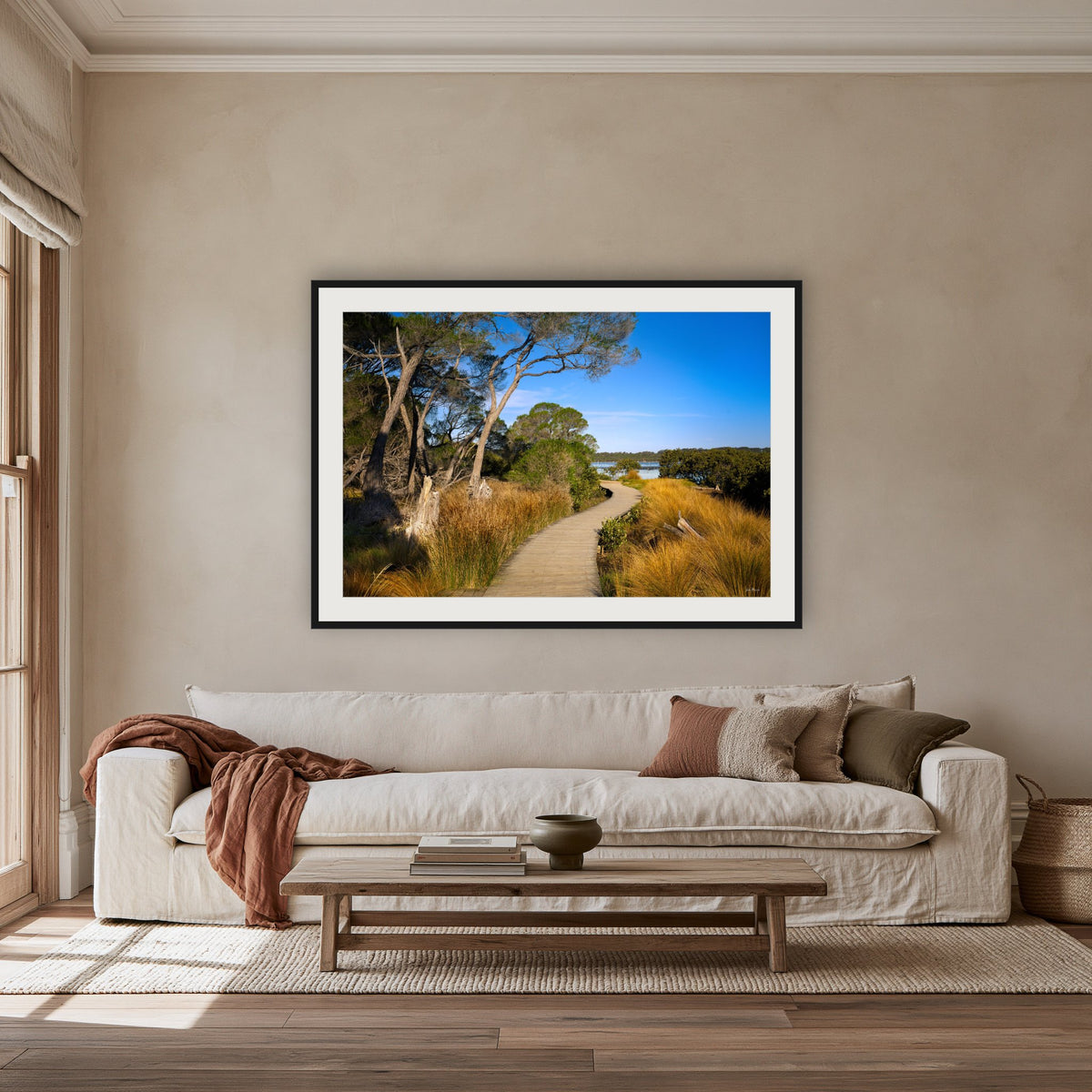 Merimbula Boardwalk through Golden Grasses framed print in black with a white mat, above an oatmeal linen sofa in a lounge room