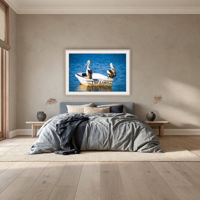 Pelican's Limo framed print in oak with a white mat, above a dusty blue linen bed in a bedroom