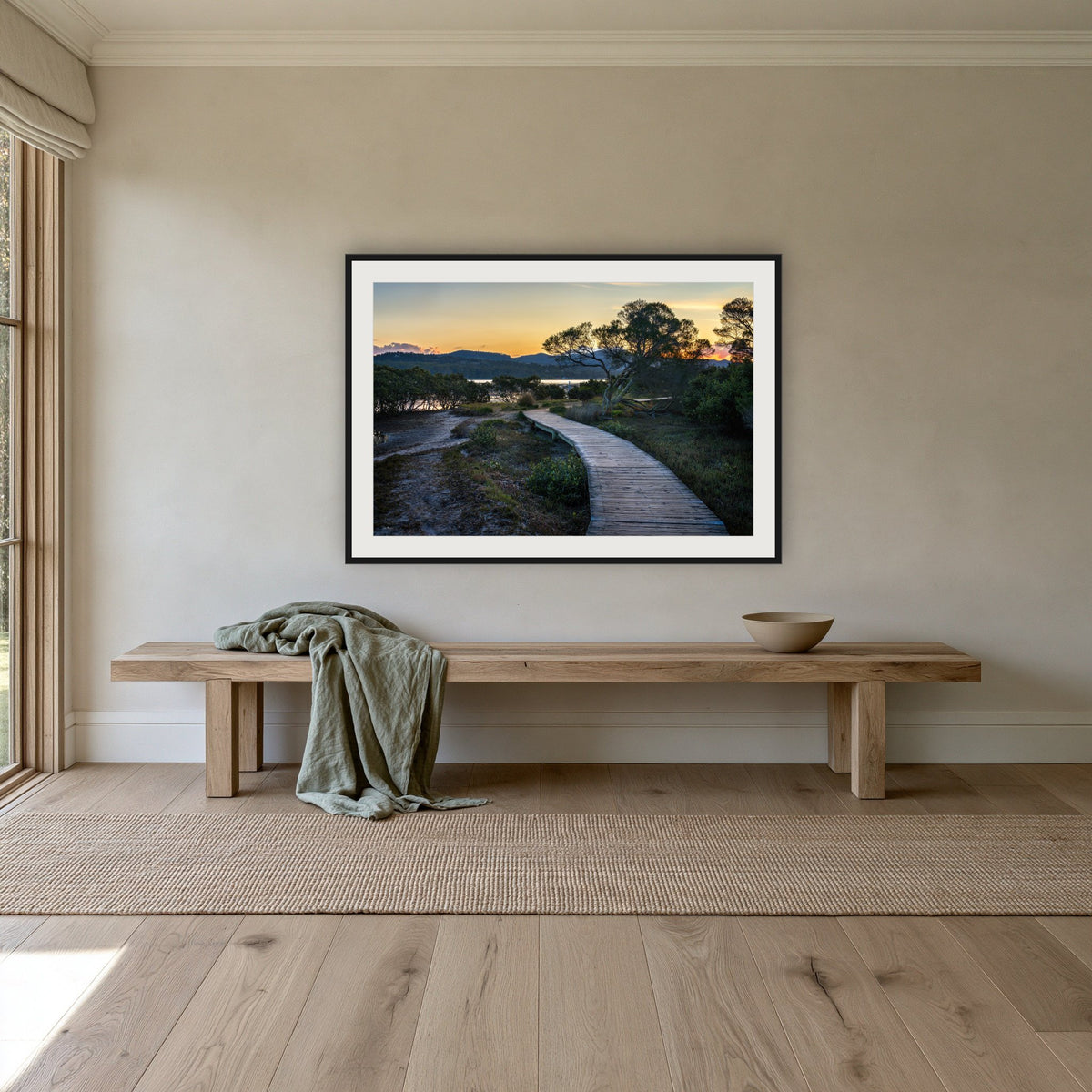 Merimbula Boardwalk at Dusk framed print in black with a white mat, above an oak bench in a hallway