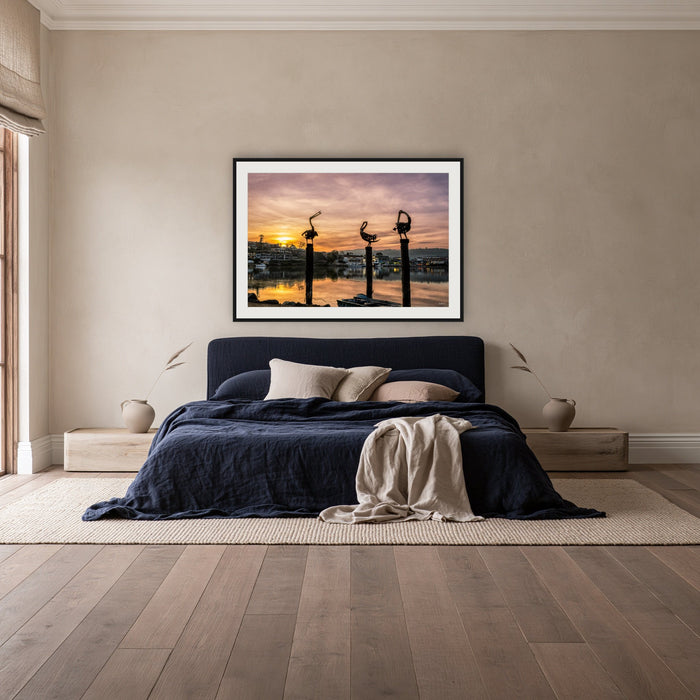 Pelican Sculptures framed print in black with a white mat, above a navy linen bed in a bedroom