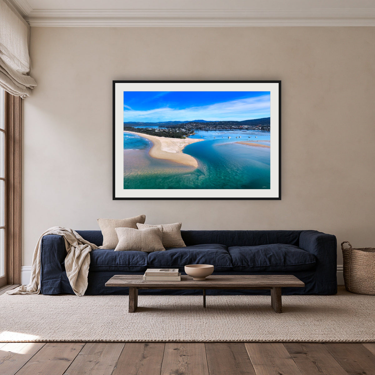 Merimbula Lake and Coastline framed print in black with a white mat, above a navy linen sofa in a lounge room