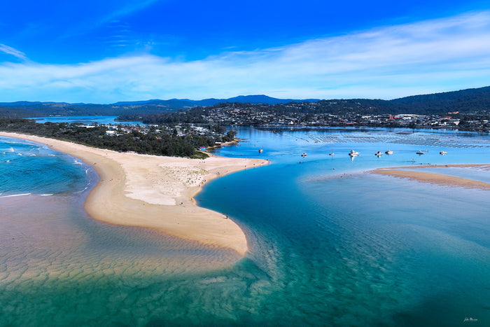 Aerial view over the Merimbula Lake sandbar in bright daylight, a turquoise channel curving between rippled sandbanks with the township and ranges beyond — fine art print