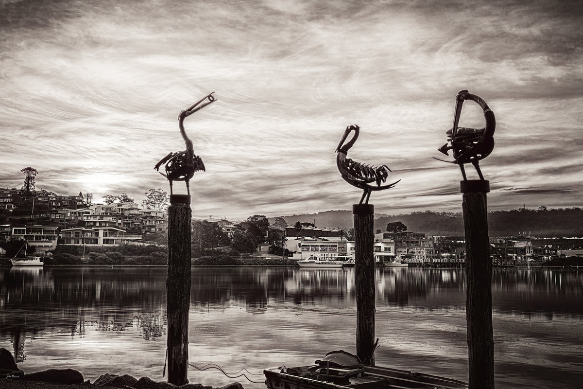 The three pelican sculptures on their pilings at Fishpen rendered in black and white, the township and lake beyond beneath a textured sky — fine art print
