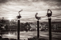 The three pelican sculptures on their pilings at Fishpen rendered in black and white, the township and lake beyond beneath a textured sky — fine art print