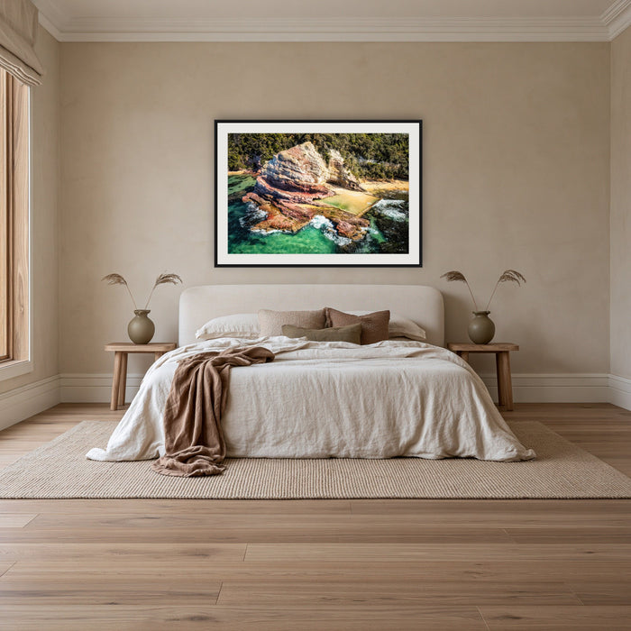 Aslings Beach Rock Pool framed print in black with a white mat, above an ivory linen bed in a bedroom