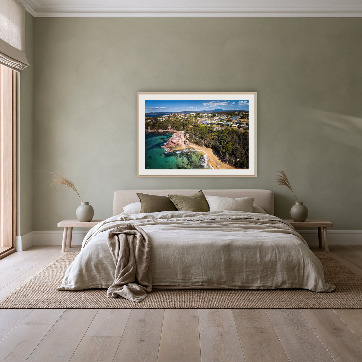 Aslings Beach from the Air framed print in oak with a white mat, above a linen bed against a sage wall in a bedroom