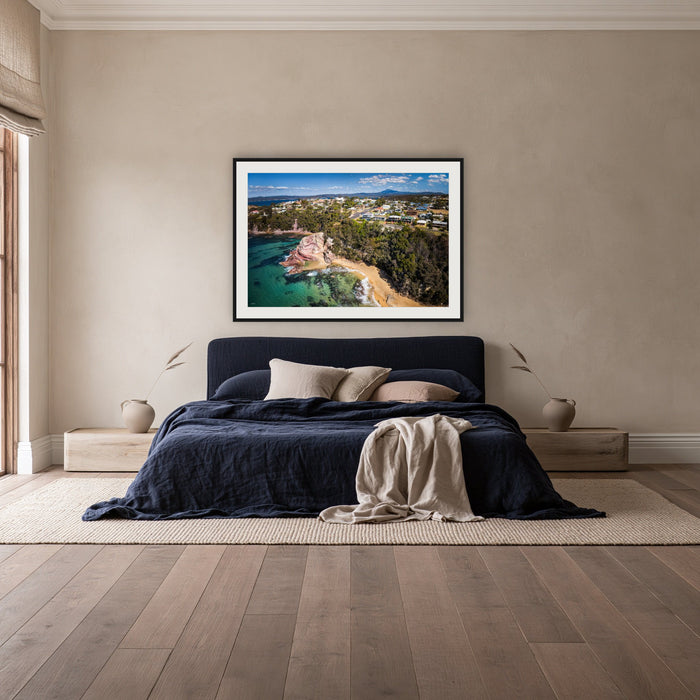Aslings Beach from the Air framed print in black with a white mat, above a navy linen bed in a bedroom