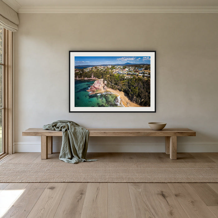 Aslings Beach from the Air framed print in black with a white mat, above an oak bench in a hallway