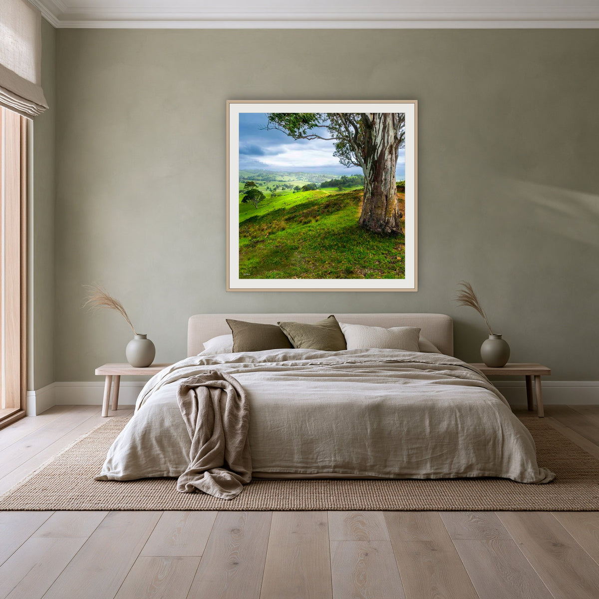 Bega Valley Countryside square framed print in oak with a white mat, above a linen bed against a sage wall in a bedroom