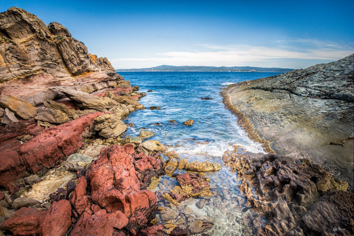 A channel of blue water between red and ochre rocks at North Head, Merimbula, a distant headland beyond — fine art print