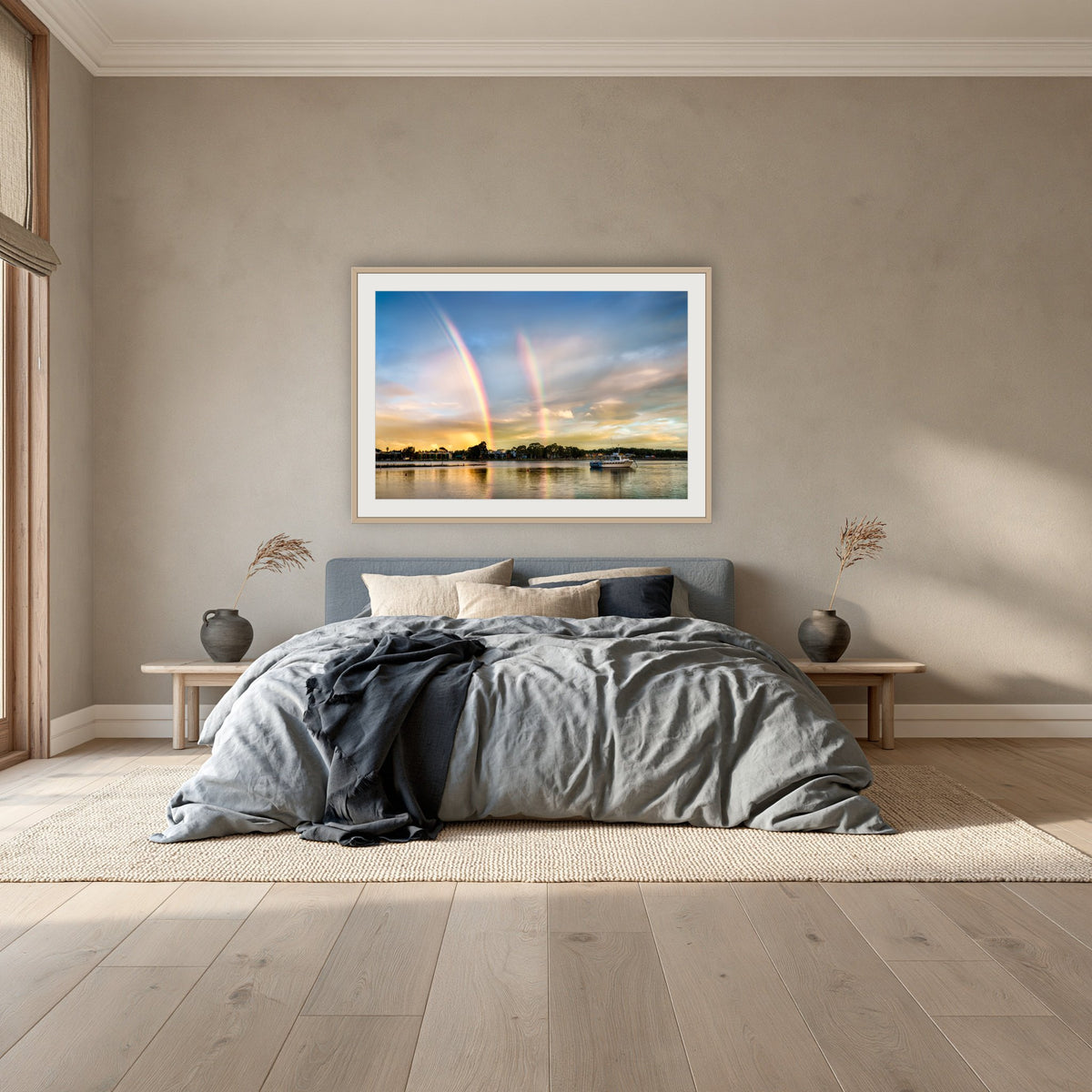 Rainbow over Merimbula Lake framed print in oak with a white mat, above a dusty blue linen bed in a bedroom