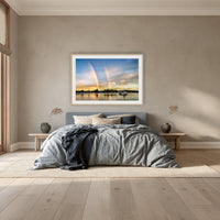 Rainbow over Merimbula Lake framed print in oak with a white mat, above a dusty blue linen bed in a bedroom