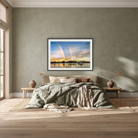 Rainbow over Merimbula Lake framed print in black with a white mat, above a khaki linen bed against a green-grey wall in a bedroom