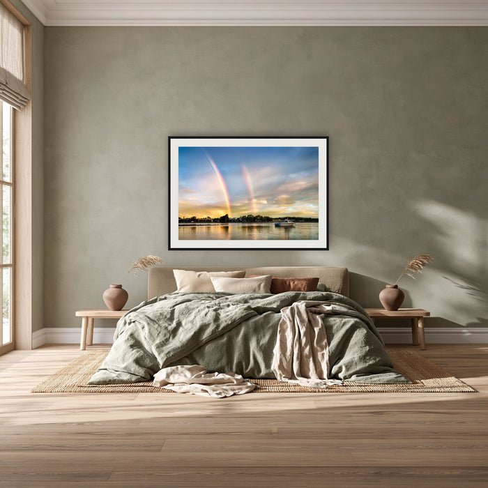 Rainbow over Merimbula Lake framed print in black with a white mat, above a khaki linen bed against a green-grey wall in a bedroom