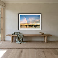 Rainbow over Merimbula Lake framed print in black with a white mat, above an oak bench in a hallway
