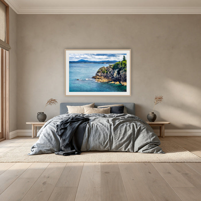 Twofold Bay Rocky Cliffs & Mt Imlay framed print in oak with a white mat, above a dusty blue linen bed in a bedroom