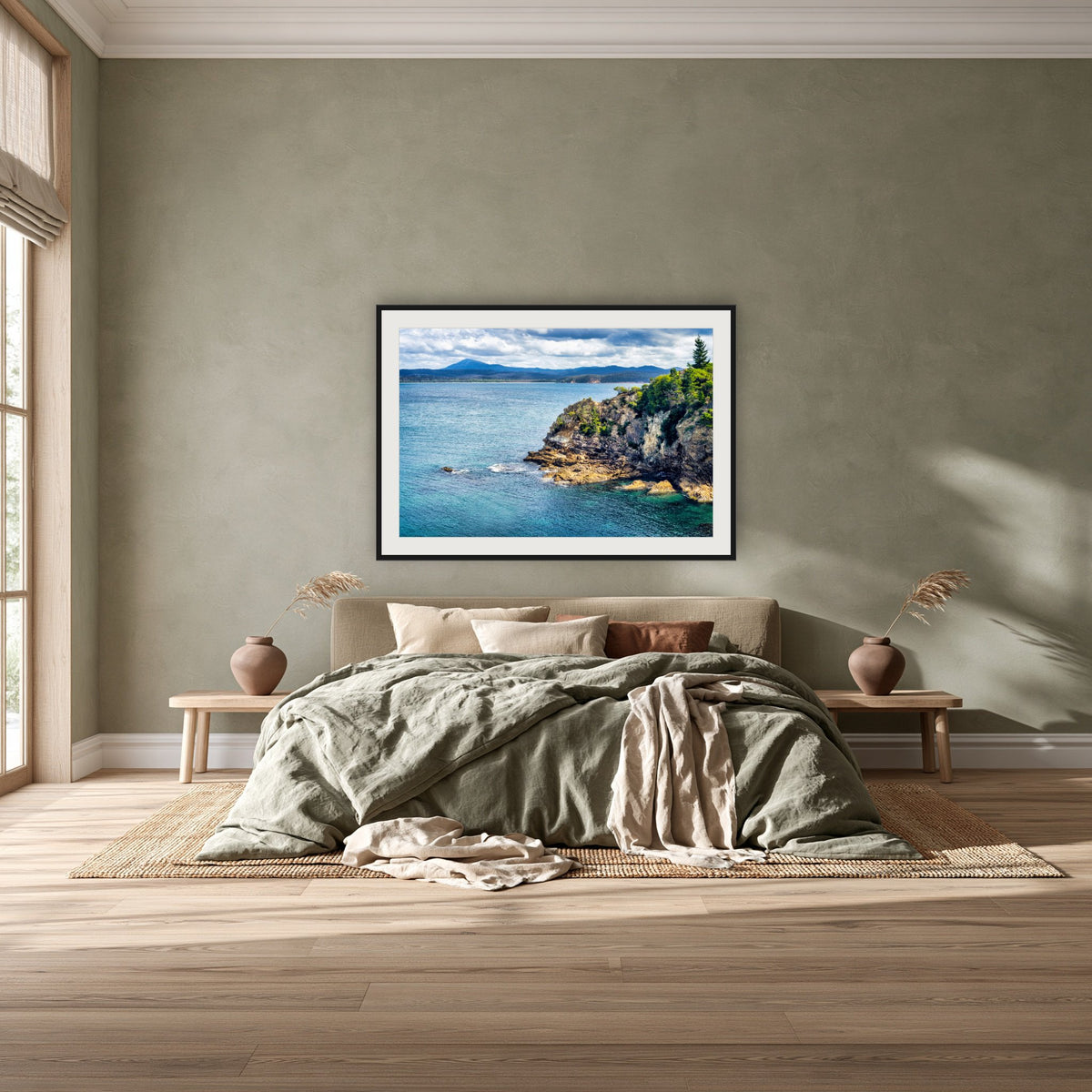 Twofold Bay Rocky Cliffs & Mt Imlay framed print in black with a white mat, above a khaki linen bed against a green-grey wall in a bedroom