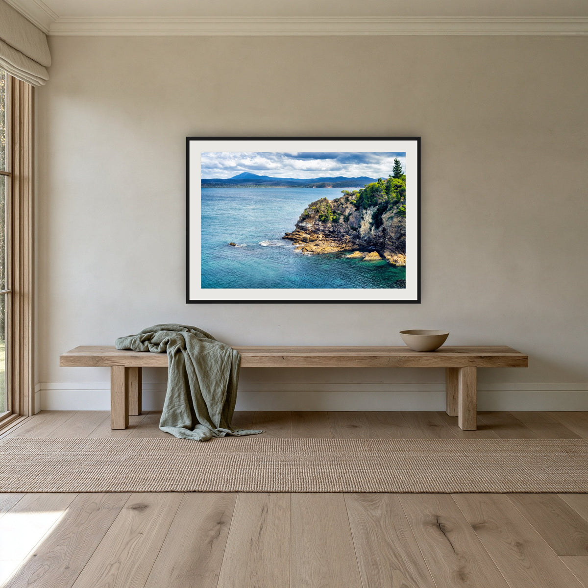 Twofold Bay Rocky Cliffs & Mt Imlay framed print in black with a white mat, above an oak bench in a hallway