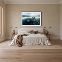 Little Boat at Fishpen Jetty framed print in black with a white mat, above an ivory linen bed in a bedroom