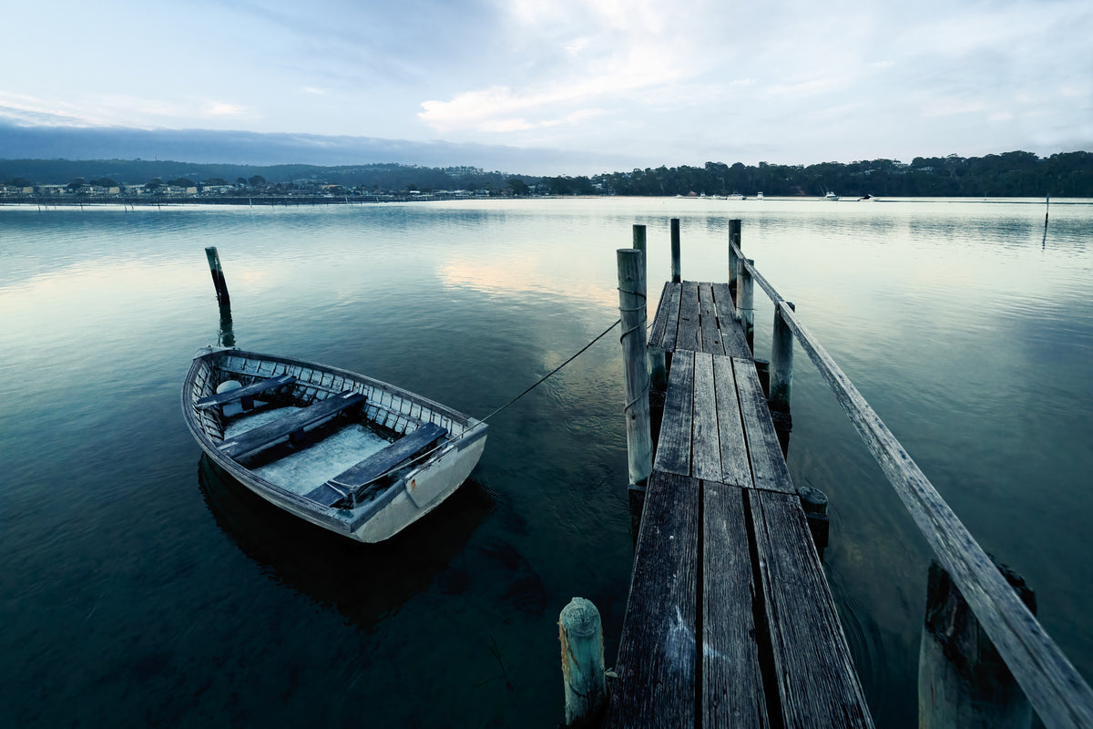 A wooden dinghy moored beside a weathered jetty on flat calm water, Merimbula Lake township across the water — fine art print
