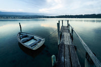 A wooden dinghy moored beside a weathered jetty on flat calm water, Merimbula Lake township across the water — fine art print