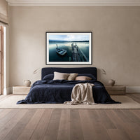 Little Boat at Fishpen Jetty framed print in black with a white mat, above a navy linen bed in a bedroom