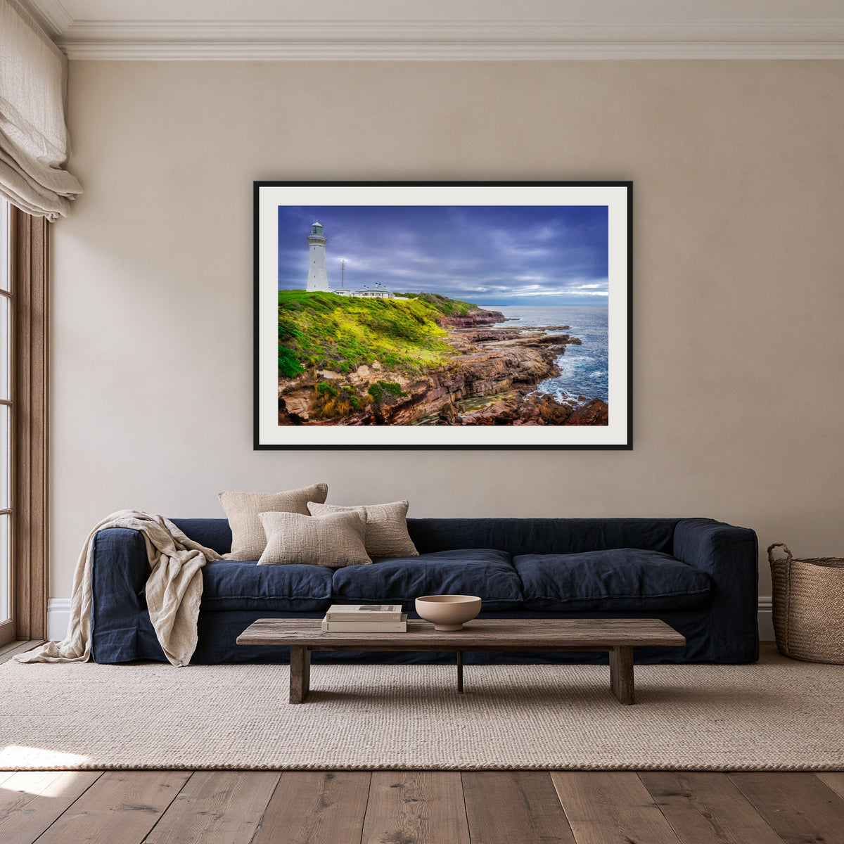 Green Cape Lighthouse framed print in black with a white mat, above a navy linen sofa in a lounge room