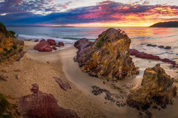 A glorious sunrise over Pambula Beach, magenta and violet cloud breaking to gold along the horizon, the light catching a tall rock outcrop while a channel of wet sand curves out to the sea — fine art print
