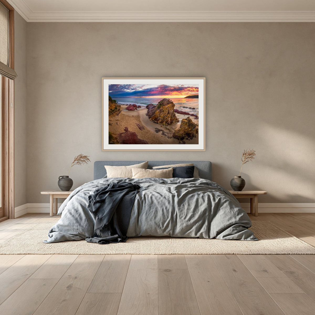 Pambula Beach Sunrise framed print in oak with a white mat, above a dusty blue linen bed in a bedroom