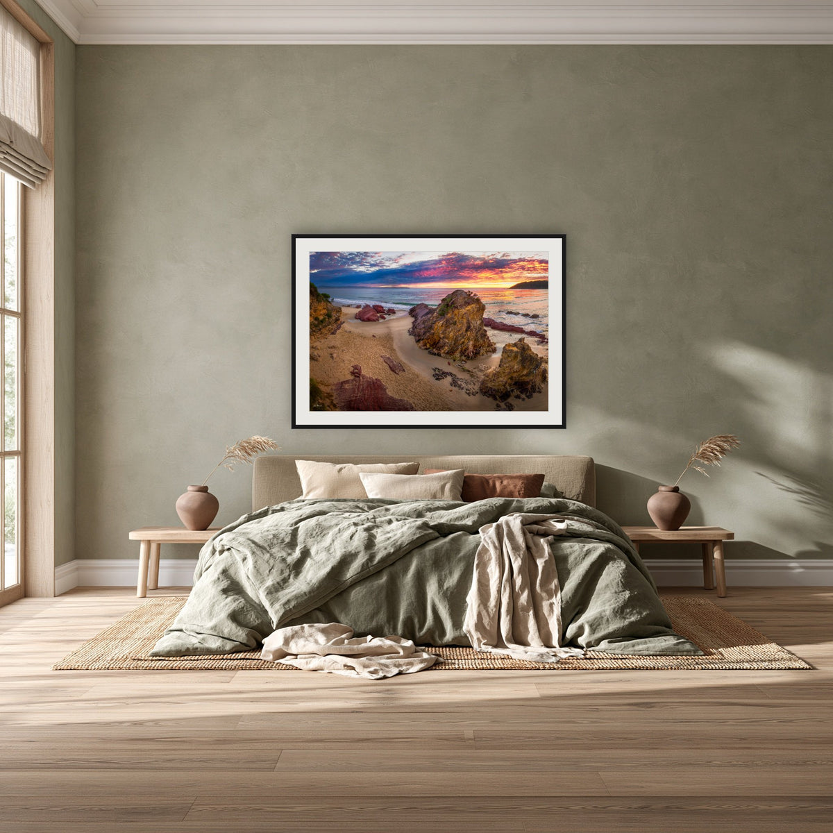 Pambula Beach Sunrise framed print in black with a white mat, above a khaki linen bed against a green-grey wall in a bedroom