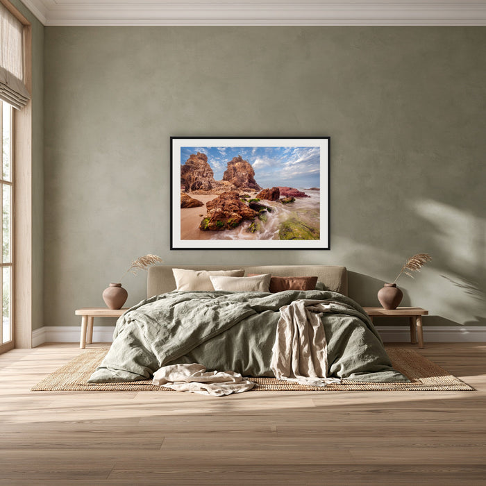 Pambula Rocks framed print in black with a white mat, above a khaki linen bed against a green-grey wall in a bedroom