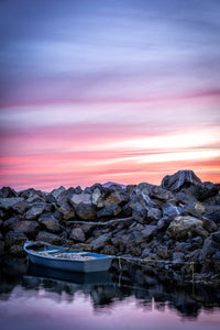 A small blue dinghy moored against a rock wall under a streaked pink and violet sunset sky, its reflection in the still water below — fine art print