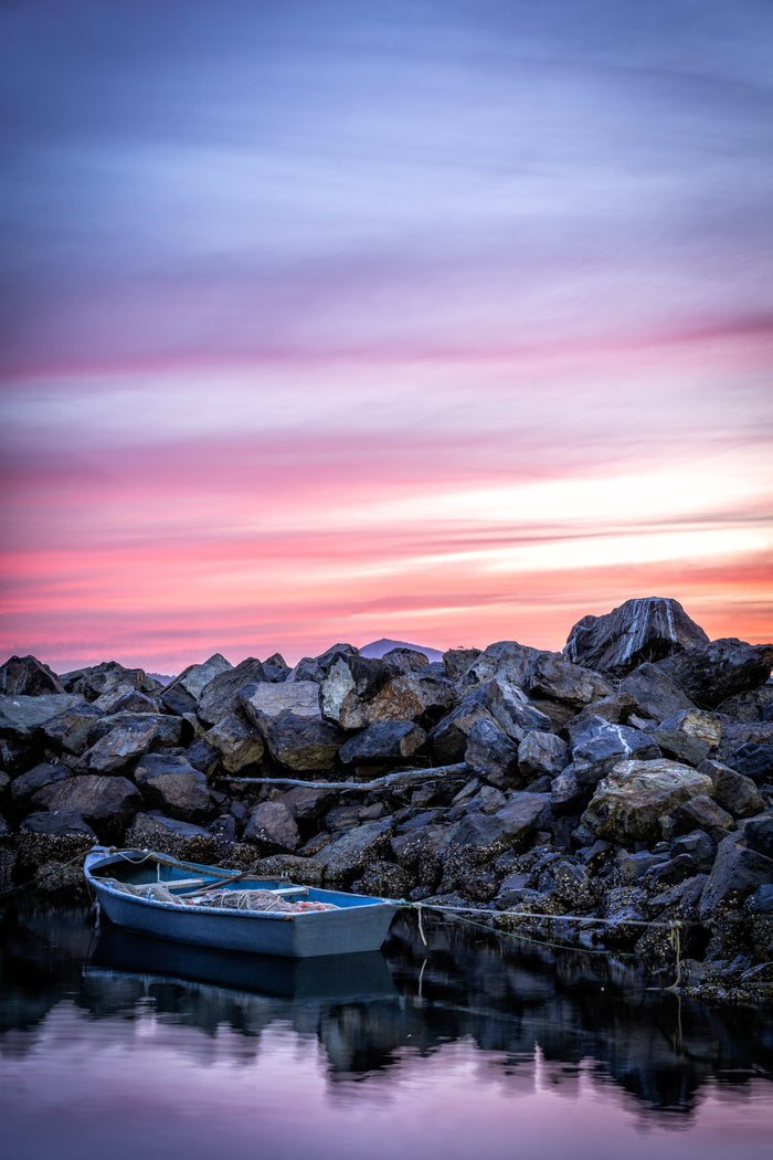 A small blue dinghy moored against a rock wall under a streaked pink and violet sunset sky, its reflection in the still water below — fine art print