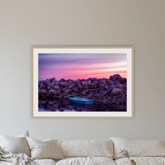 Eden Twofold Hush wall art — a lone blue dinghy against the Eden breakwall at last light, shown in an oak frame in a lounge room