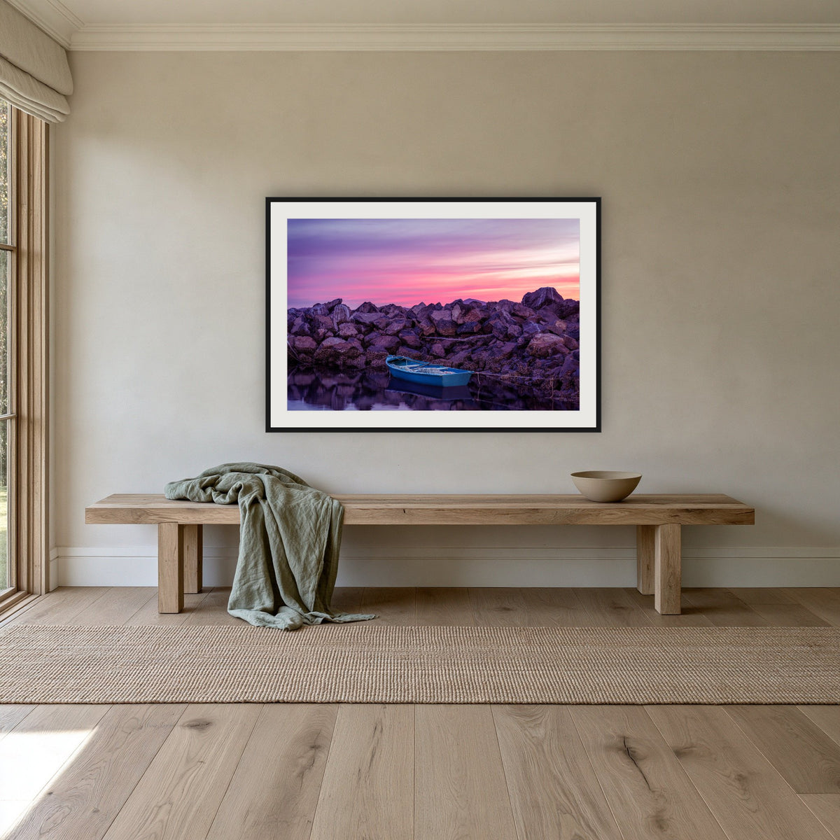 Eden Twofold Hush wall art — a lone blue dinghy against the Eden breakwall at last light, shown in a black frame with a white mat above an oak bench in a hallway