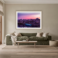 Eden Twofold Hush wall art — a lone blue dinghy against the Eden breakwall at last light, shown in an oak frame with a white mat above an olive linen sofa in a lounge room