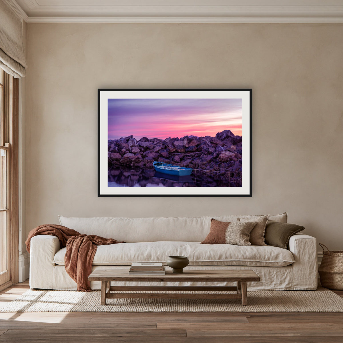 Eden Twofold Hush wall art — a lone blue dinghy against the Eden breakwall at last light, shown in a black frame with a white mat above an oatmeal linen sofa in a lounge room