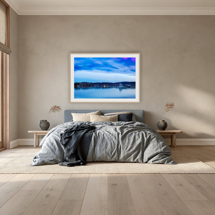 Dawn over Merimbula Lake framed print in oak with a white mat, above a dusty blue linen bed in a bedroom
