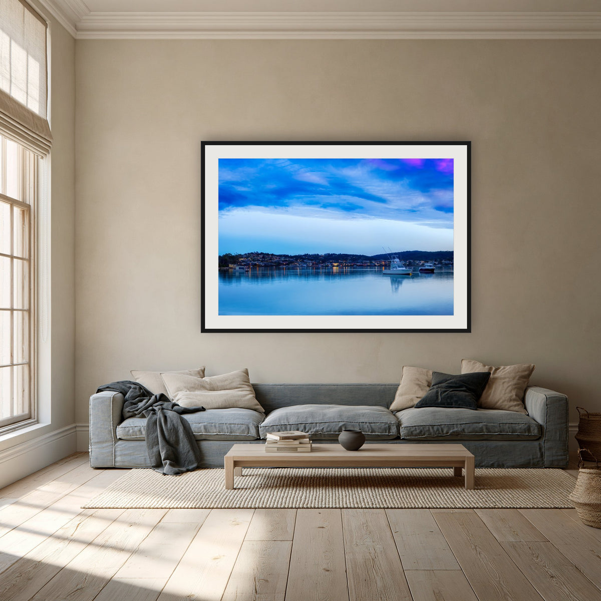 Dawn over Merimbula Lake framed print in black with a white mat, above a dusty blue linen sofa in a lounge room