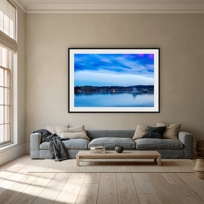 Dawn over Merimbula Lake framed print in black with a white mat, above a dusty blue linen sofa in a lounge room