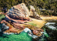 Aslings Beach Rock Pool, Eden NSW — aerial view of the ocean pool beneath a headland of layered red and cream rock, with turquoise water and surf